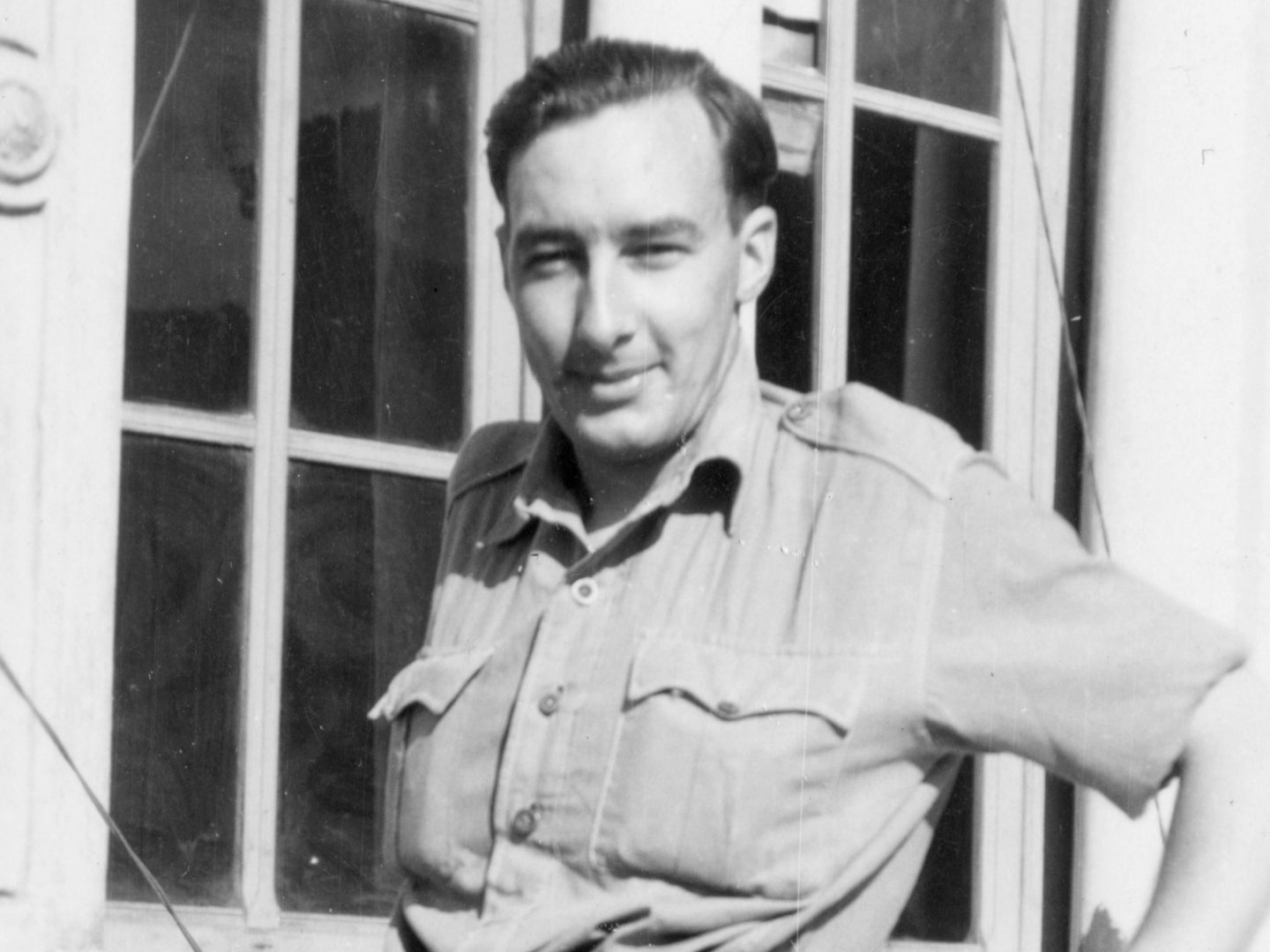 A historic black and white photo of a man in a uniform shirt. He is standing in front of a window and squinting into the sun.