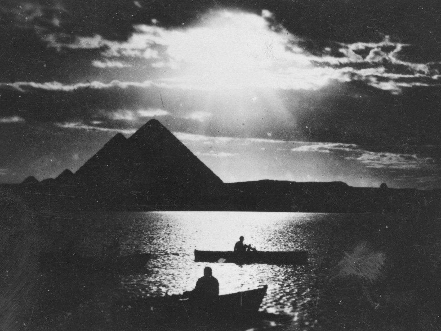 A historic black and white photo of the Pyramids of Giza. The photo is taken from the Nile straight into the sun, producing a highly contrasted image. The sun is above the pyramids, and several small boats are visible on the water in the foreground.