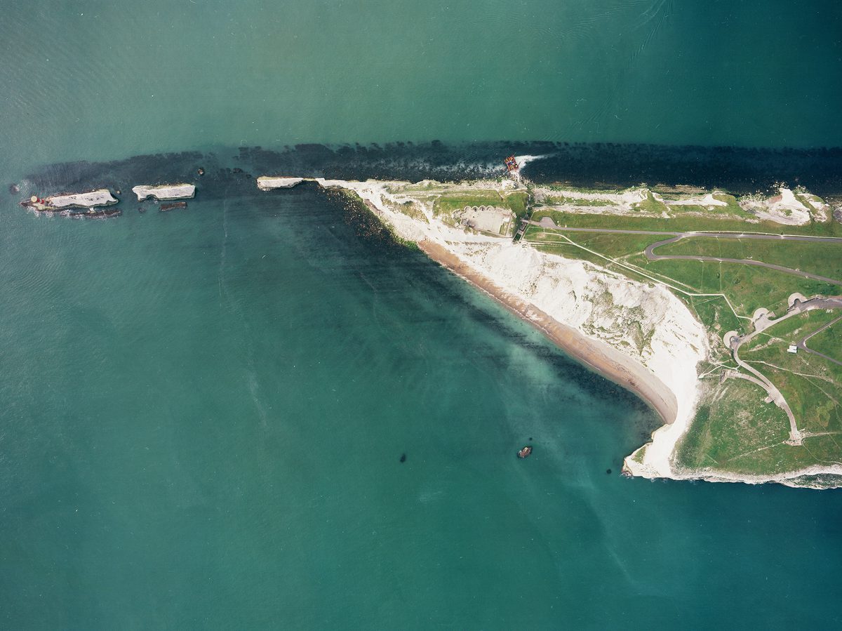 A colour aerial photo of a headland protruding into the sea from right to left. The headland is formed of white chalk cliffs and is topped with grass. It narrows to a sharp point and several cliff stacks. A lighthouse is visible on the final stack.