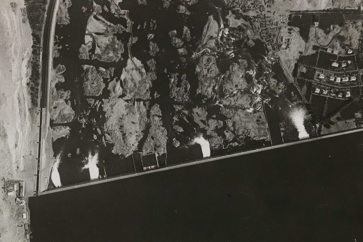 A historic black and white aerial photo of a dam. The dam runs horizontally across the image, with the water appearing very black along the lower edge of the image. Rocky land is visible in the upper half of the image, with water outflows visible as white splashes.