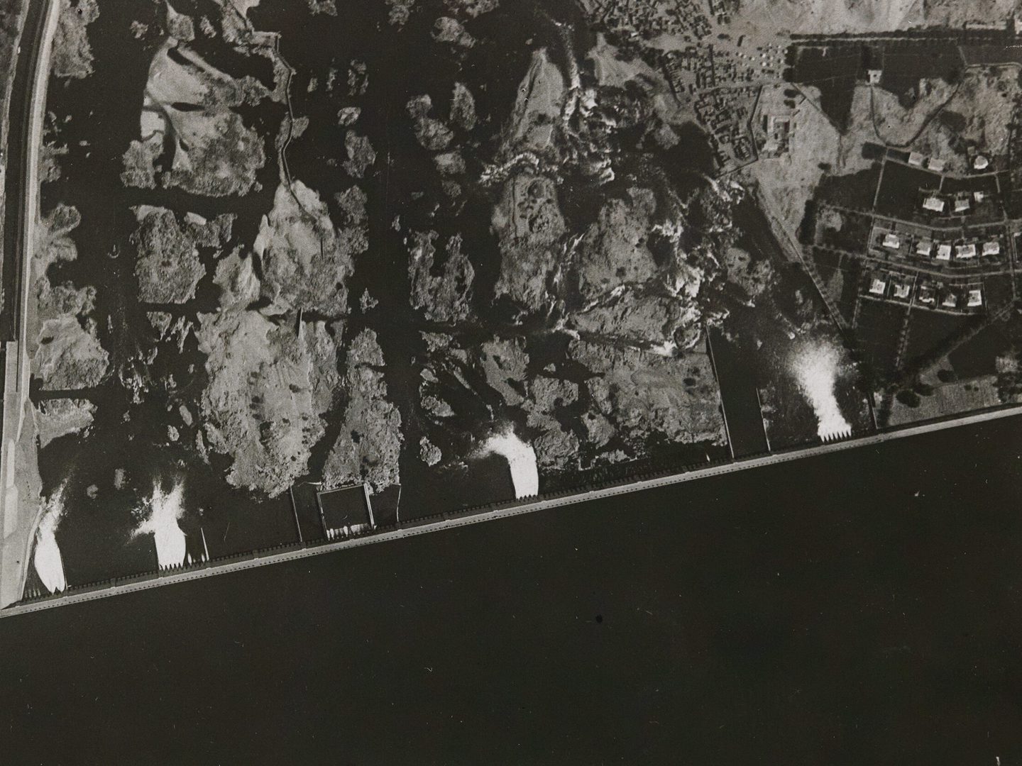 A historic black and white aerial photo of a dam. The dam runs horizontally across the image, with the water appearing very black along the lower edge of the image. Rocky land is visible in the upper half of the image, with water outflows visible as white splashes.