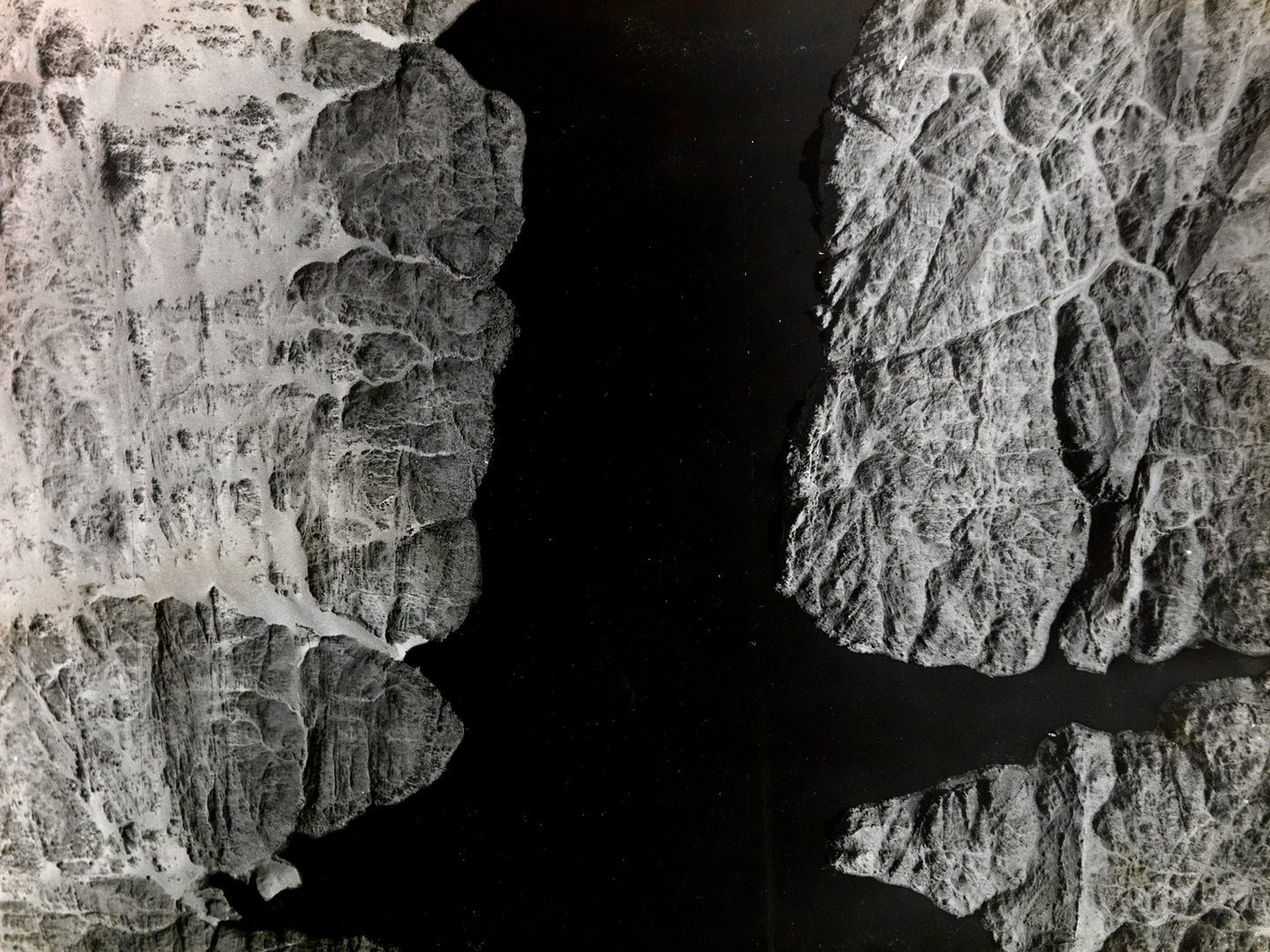 A historic black and white vertical aerial photo of a wide river with fissured rocky banks. The river runs from the top of the image to the bottom, with the banks at the right and left sides.