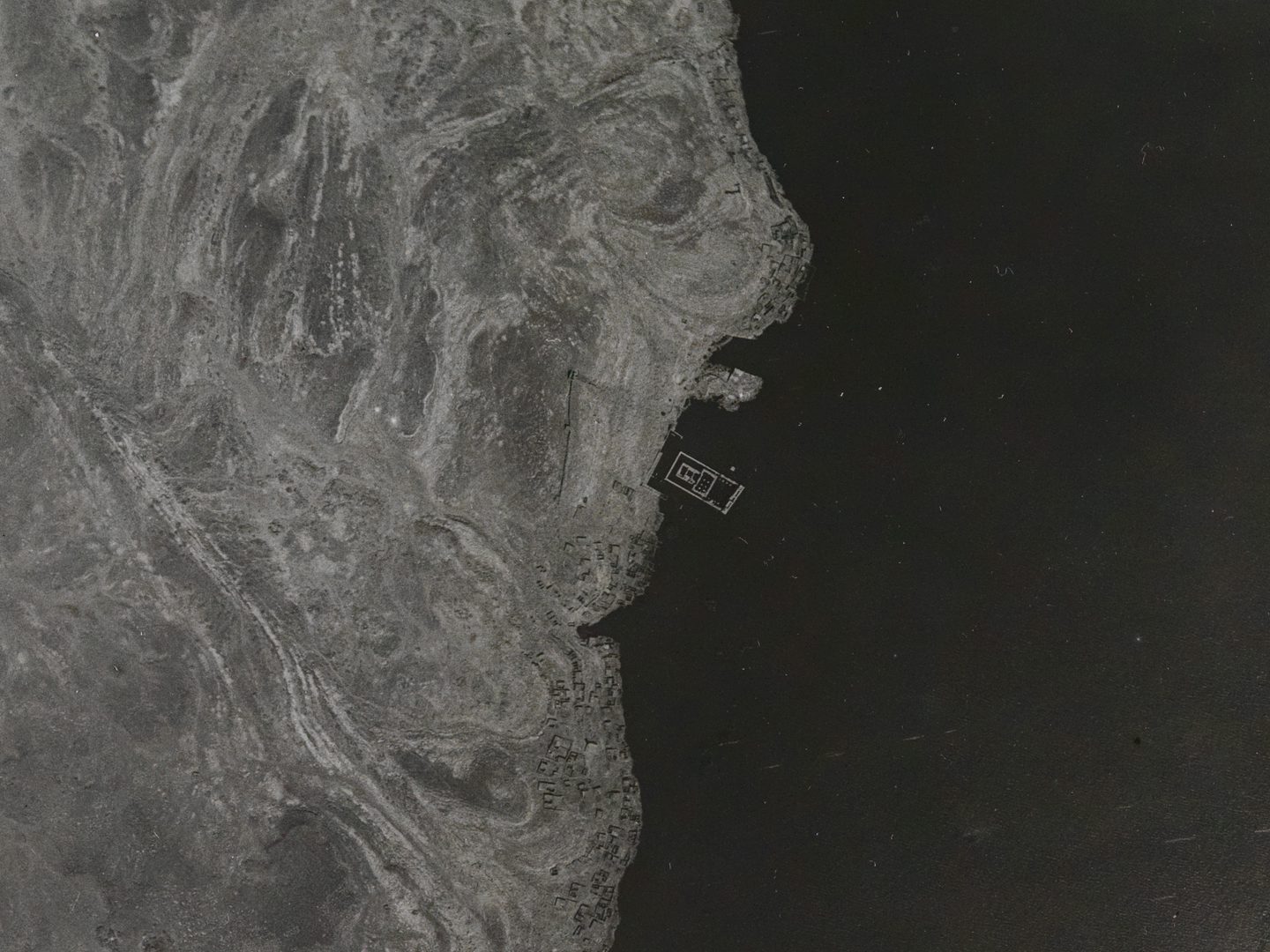 A historic black and white vertical aerial photo of a river. The river fills the right-hand side of the image, with a sandy bank filling the left-hand side. In the middle of the image, just off the bank in the water is a complex with rectangular walls.