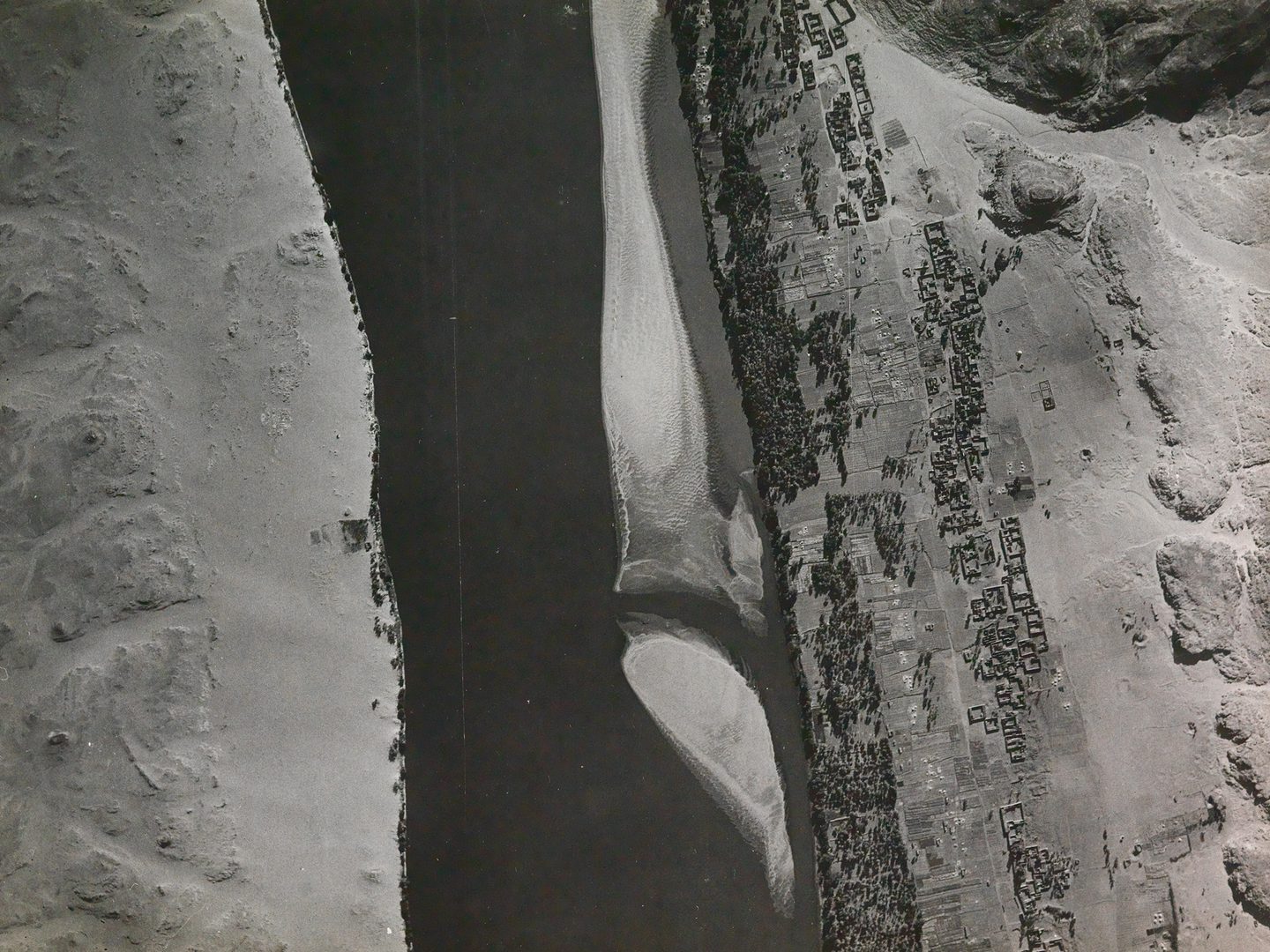 A historic black and white vertical aerial photo of a river running between sandy banks. The river runs from the top of the image to the bottom. A settlement stretches along the right-hand bank, with lines of trees by the water's edge.