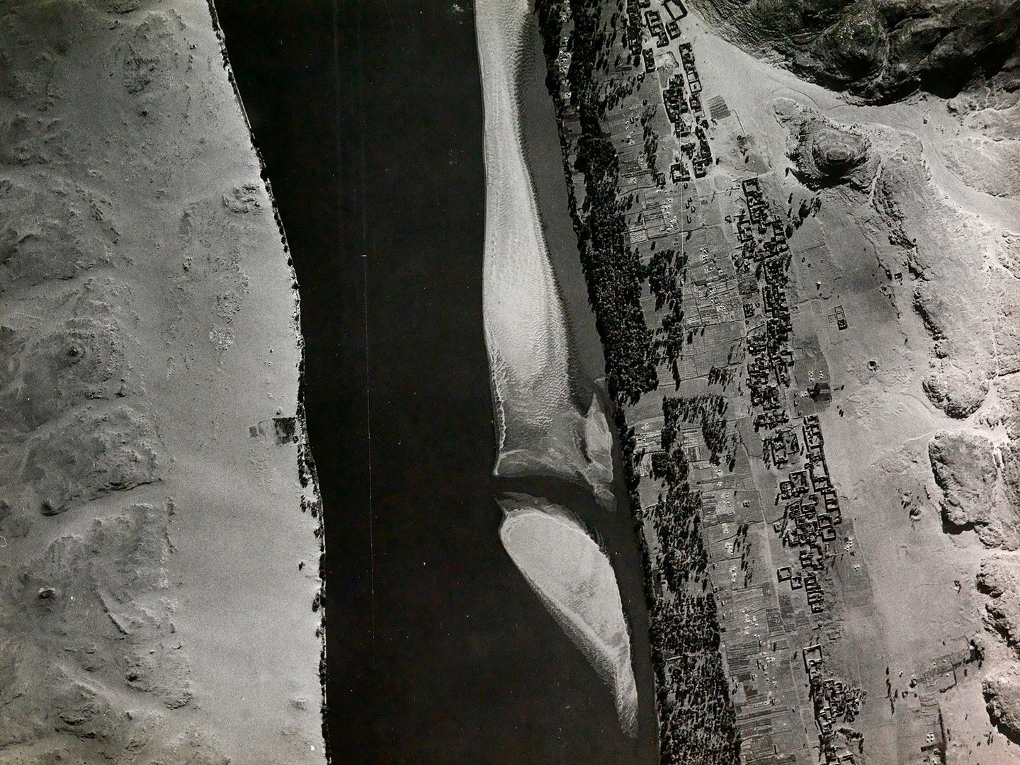 A historic black and white vertical aerial photo of a river running between sandy banks. The river runs from the top of the image to the bottom. A settlement stretches along the right-hand bank, with lines of trees by the water's edge.