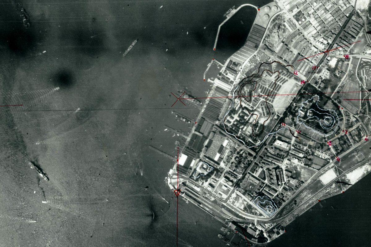 A historic black and white vertical aerial photo with the sea to the left and land to the right. There are several ships on the sea and more at piers projecting from the land. The land is heavily built-up. There are lines and annotations in red and white pencil.