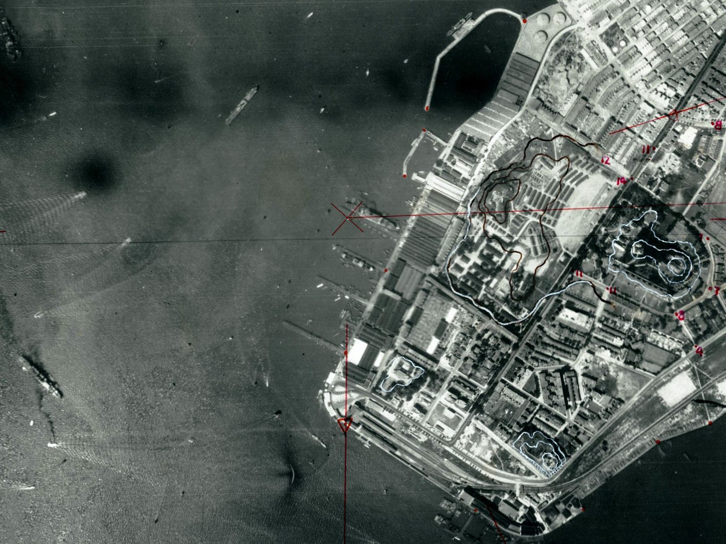 A black and white aerial photo with the sea to the left and land to the right. There are several ships on the sea and more at piers projecting from the land. The land is heavily built-up. There are lines and annotations in red and white pencil.