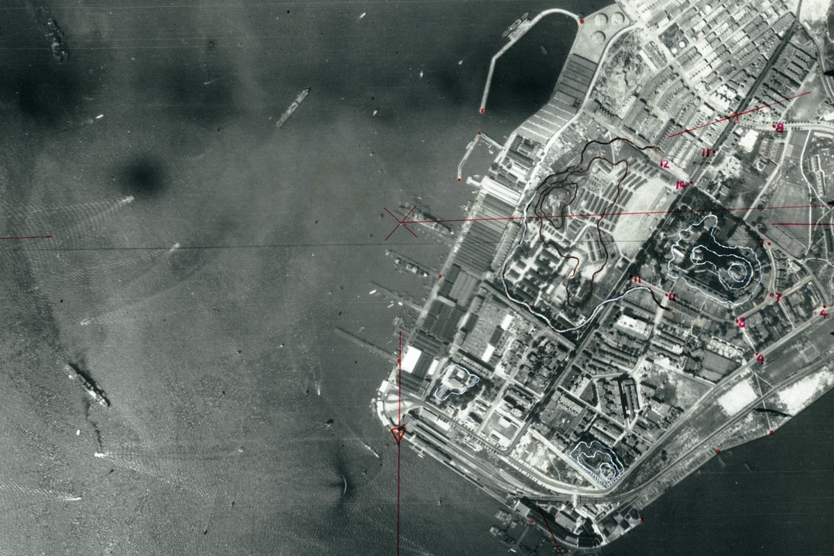 A black and white aerial photo with the sea to the left and land to the right. There are several ships on the sea and more at piers projecting from the land. The land is heavily built-up. There are lines and annotations in red and white pencil.