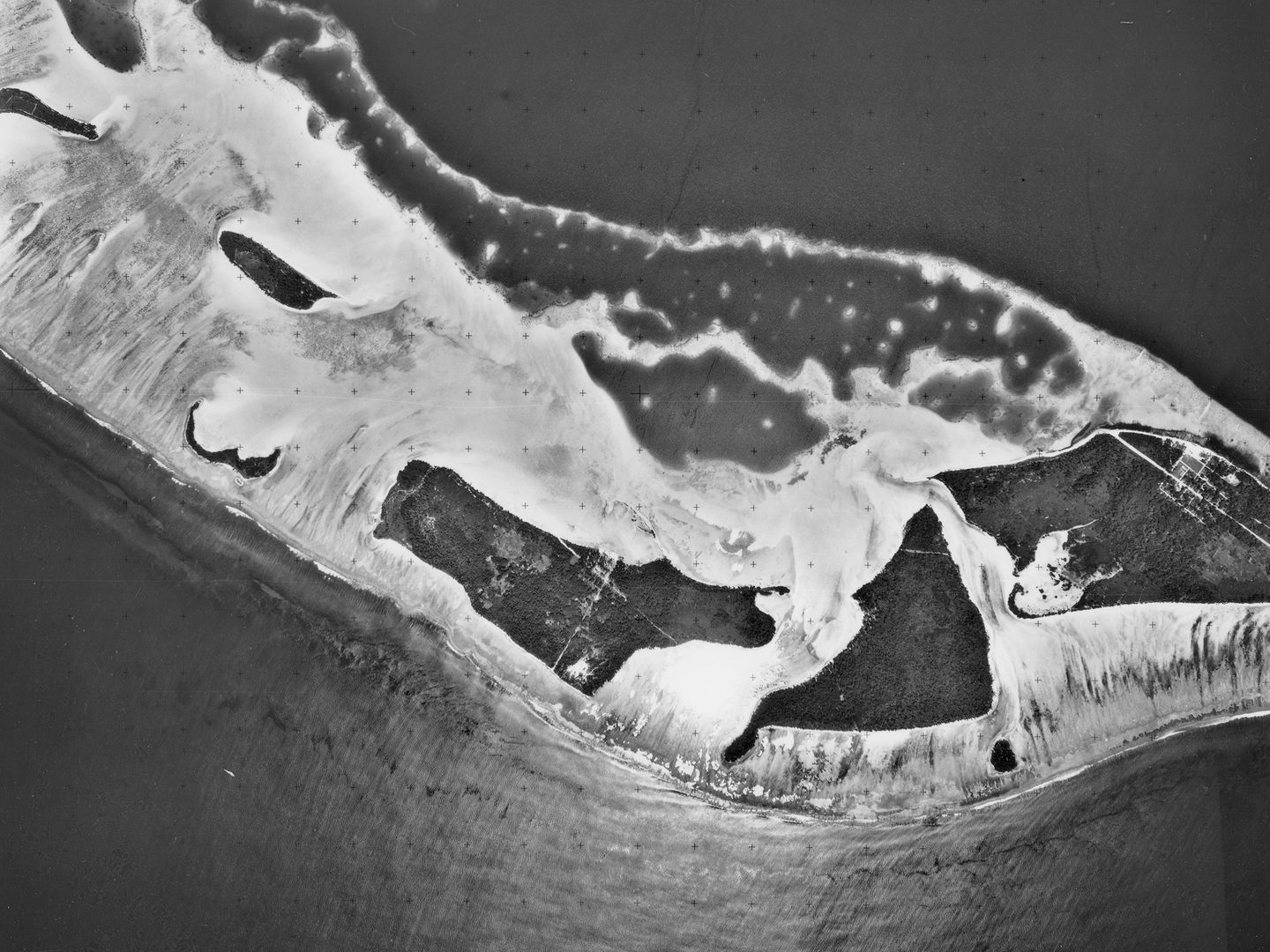 A historic black and white vertical aerial photo of several irregular small islands. The islands are surrounded by light-coloured areas of sand or coral, which in turn are surrounded by the dark sea.