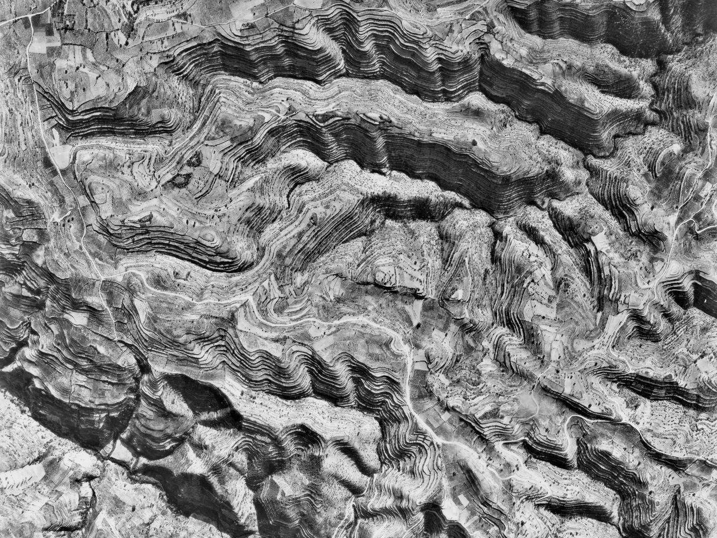 A black and white vertical aerial photo of a landscape. A complex network of hills and valleys crosses the landscape. The hills are very heavily terraced.