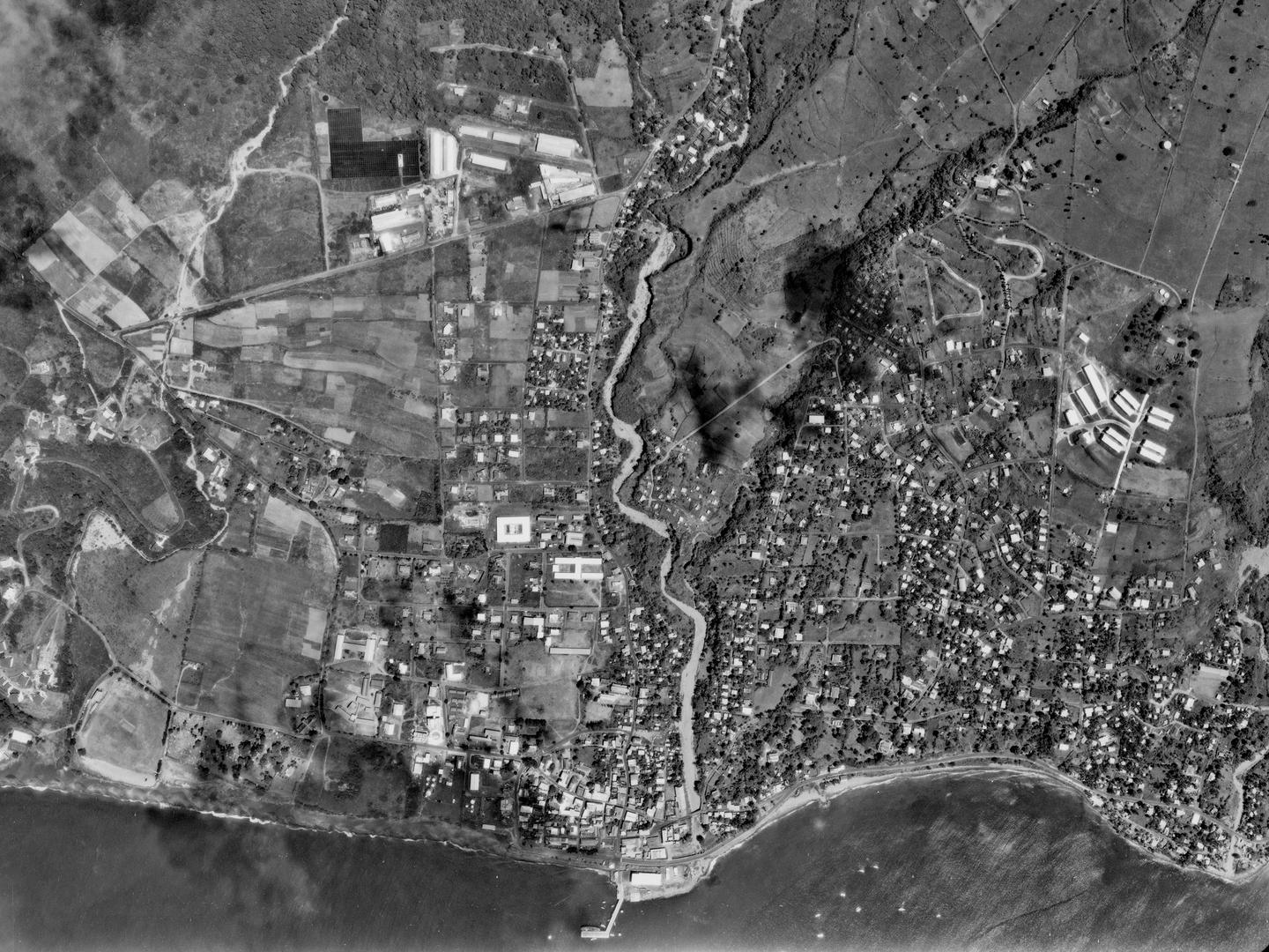 A black and white aerial photo of a town on a coastline. The town is comprised of scattered houses interspersed with fields. A river runs through the middle of the town. A small jetty protrudes into the sea.