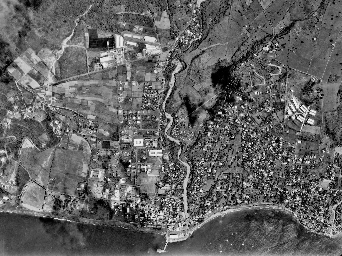 A black and white aerial photo of a town on a coastline. The town is comprised of scattered houses interspersed with fields. A river runs through the middle of the town. A small jetty protrudes into the sea.