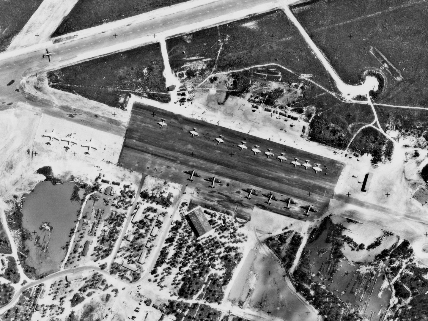 A historic black and white vertical aerial photo of an airfield. 19 aircraft are parked on a large area of concrete in the middle of the image: 3 Valiants, 6 Shackletons and 10 Canberras. Buildings among trees surround the parking area.