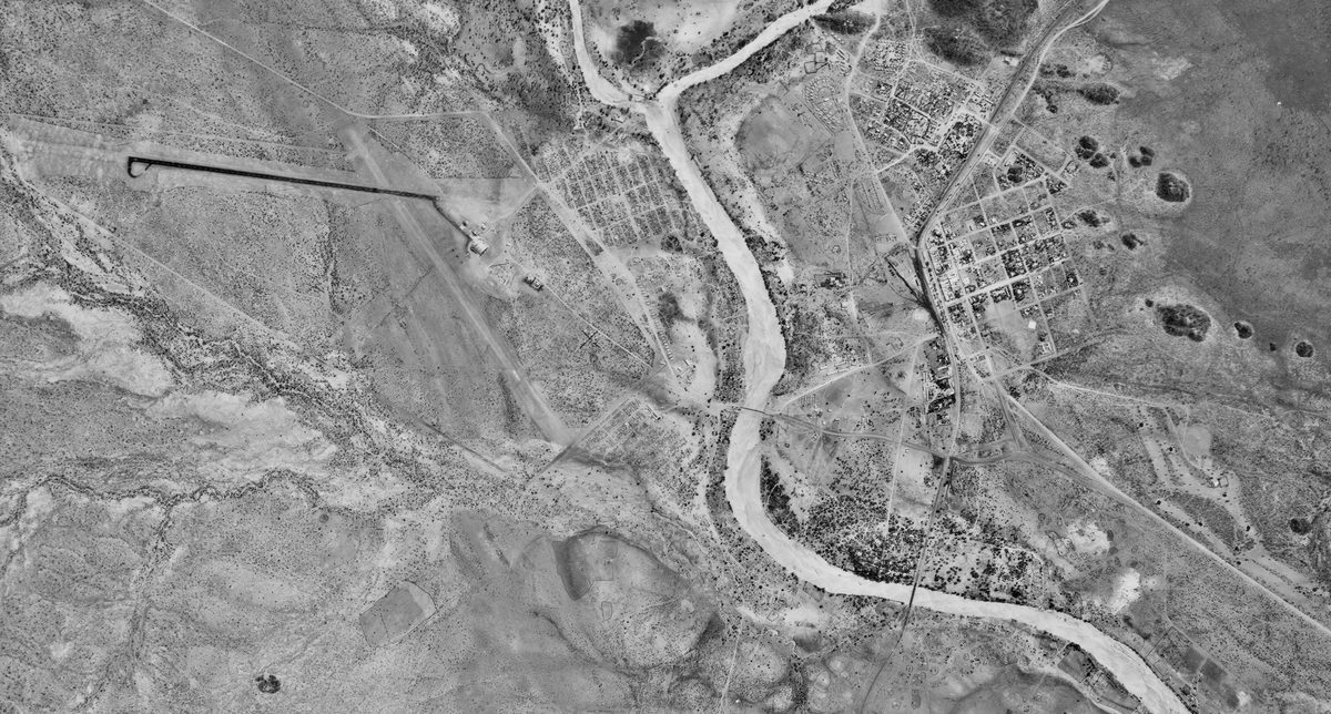 A historic black and white vertical aerial photo of a landscape with a river running through it. On the right of the river is a small settlement. To the left is an arid landscape with a few water courses running through it.