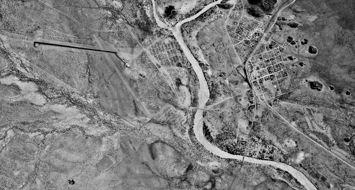 A historic black and white vertical aerial photo of a landscape with a river running through it. On the right of the river is a small settlement. To the left is an arid landscape with a few water courses running through it.