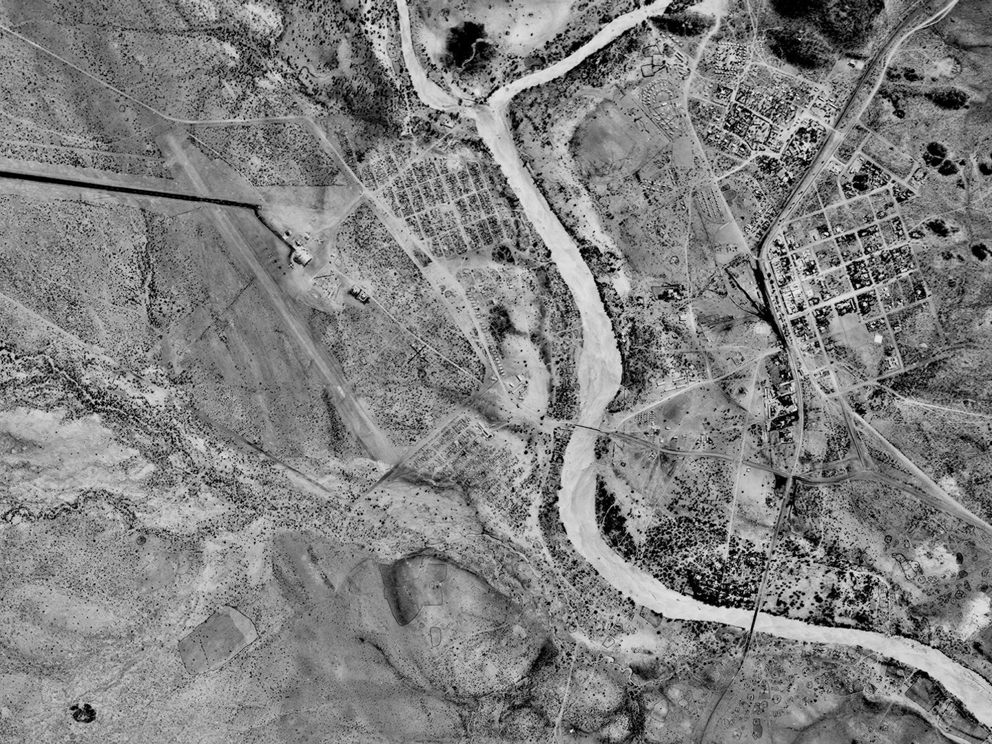 A historic black and white vertical aerial photo of a landscape with a river running through it. On the right of the river is a small settlement. To the left is an arid landscape with a few water courses running through it.