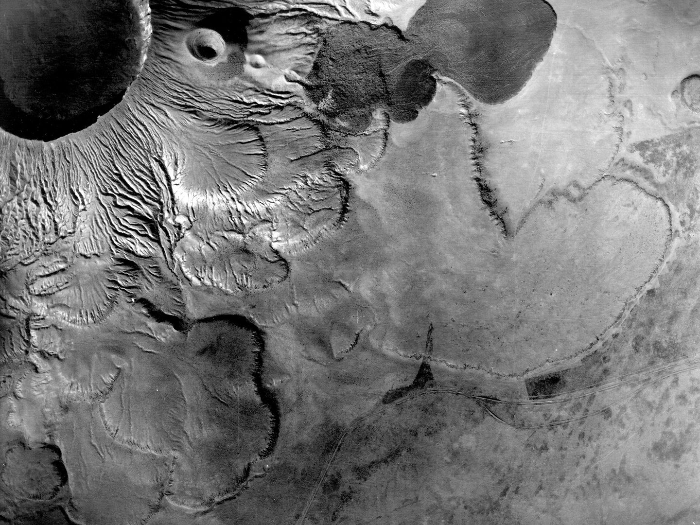 A black and white vertical aerial photo. A large volcanic crater occupies the top left corner, with fissured slopes running down to a plain below.
