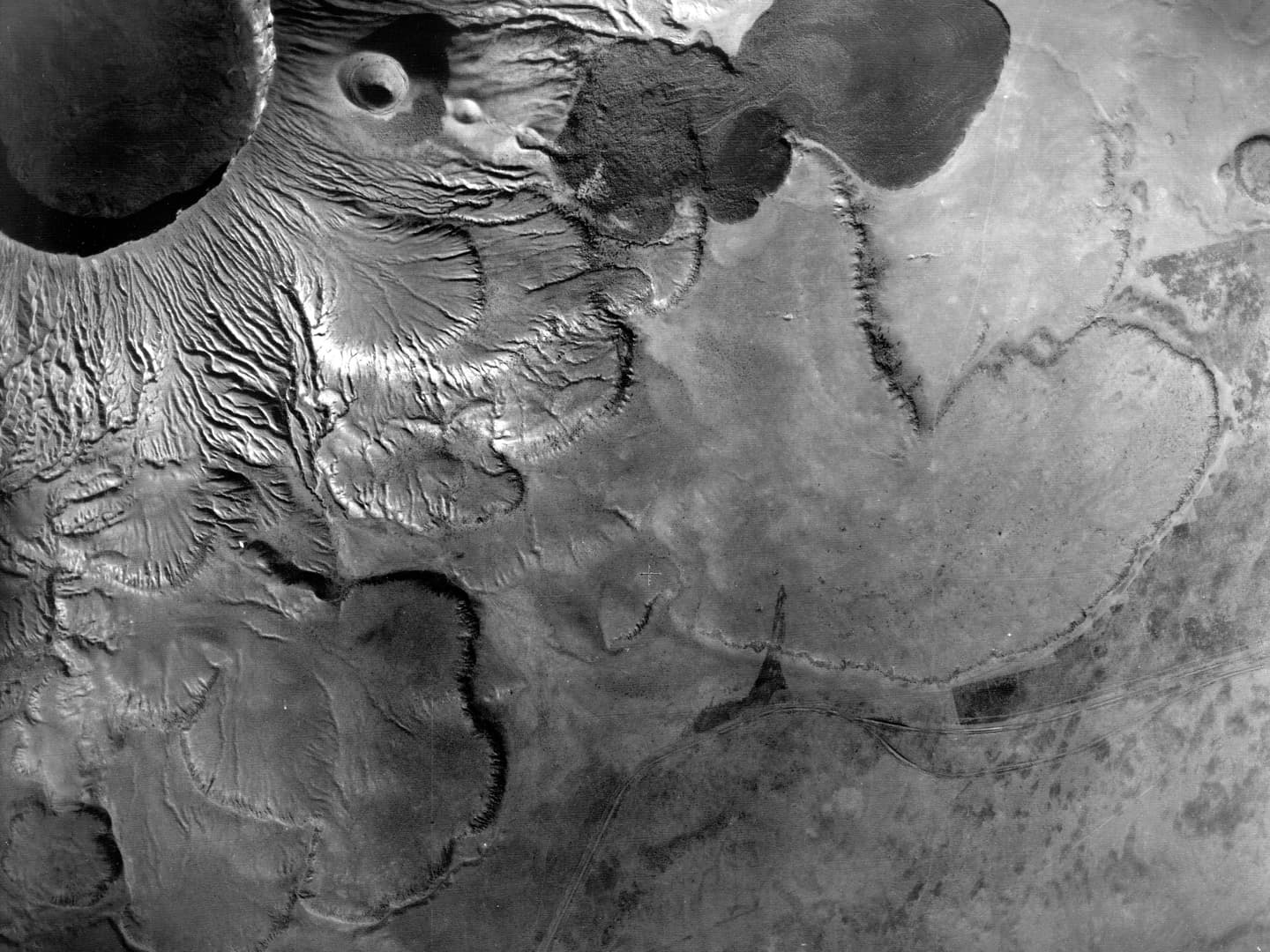 A black and white vertical aerial photo. A large volcanic crater occupies the top left corner, with fissured slopes running down to a plain below.