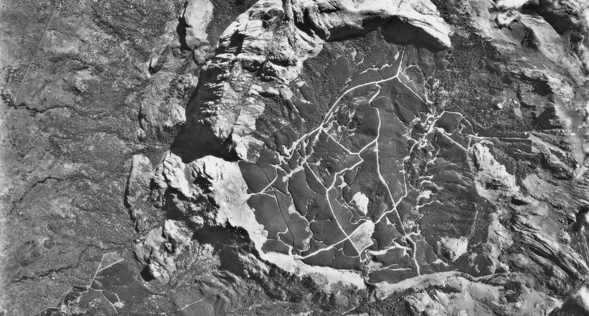 A historic black and white vertical aerial photo of mountainous terrain. A semi-circle of rock cuts through the middle of the image, forming a crater in which trails or paths are visible.