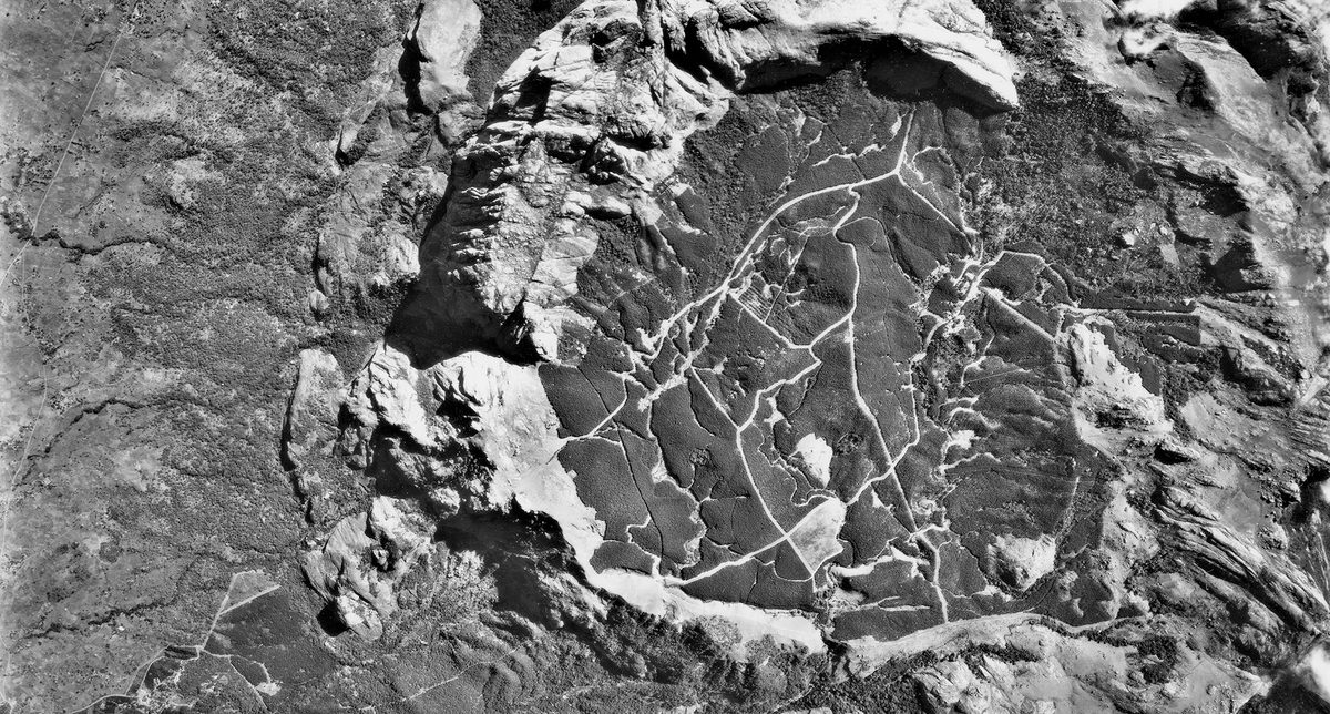 A historic black and white vertical aerial photo of mountainous terrain. A semi-circle of rock cuts through the middle of the image, forming a crater in which trails or paths are visible.