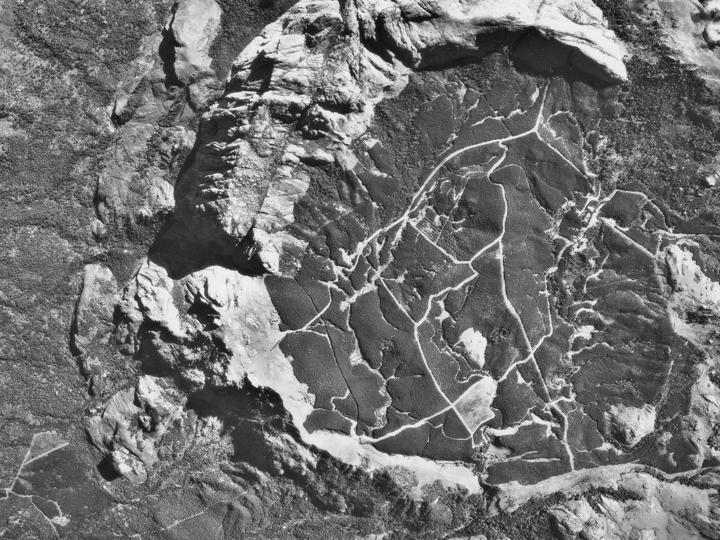 A historic black and white vertical aerial photo of mountainous terrain. A semi-circle of rock cuts through the middle of the image, forming a crater in which trails or paths are visible.