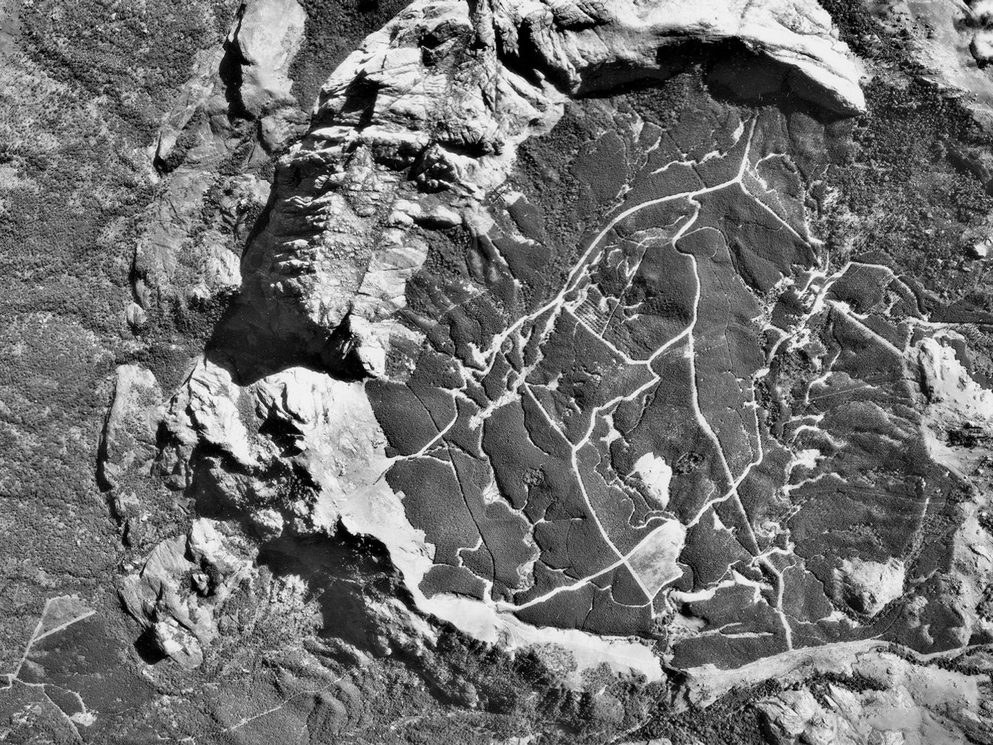 A historic black and white vertical aerial photo of mountainous terrain. A semi-circle of rock cuts through the middle of the image, forming a crater in which trails or paths are visible.