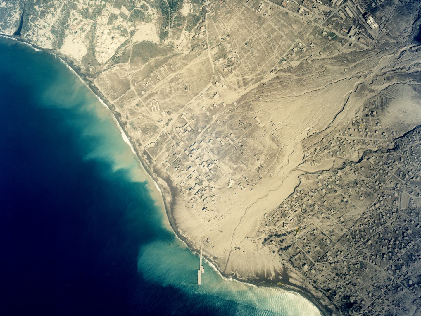 A colour aerial photo of the same town on the coast as before. It is now covered in yellow dust. The houses are buried and the river is choked with ash. The dust has spilled into the sea, staining the blue water yellow along the coast.