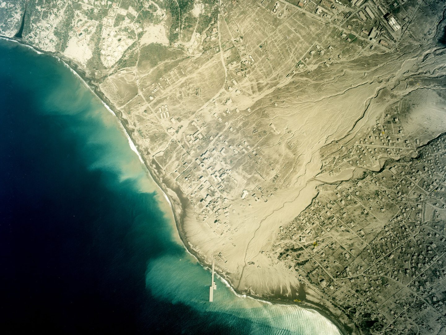 A colour aerial photo of the same town on the coast as before. It is now covered in yellow dust. The houses are buried and the river is choked with ash. The dust has spilled into the sea, staining the blue water yellow along the coast.