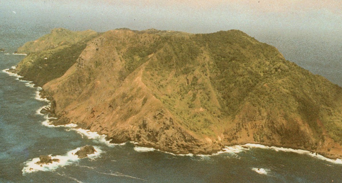 A historic colour oblique aerial photo of Pitcairn - a green island surrounded by a blue ocean and fringed by white waves.