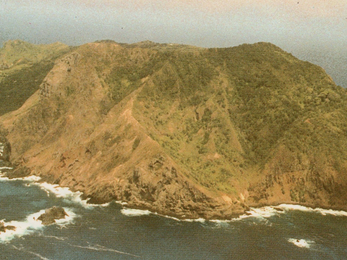 A historic colour oblique aerial photo of Pitcairn - a green island surrounded by a blue ocean and fringed by white waves.