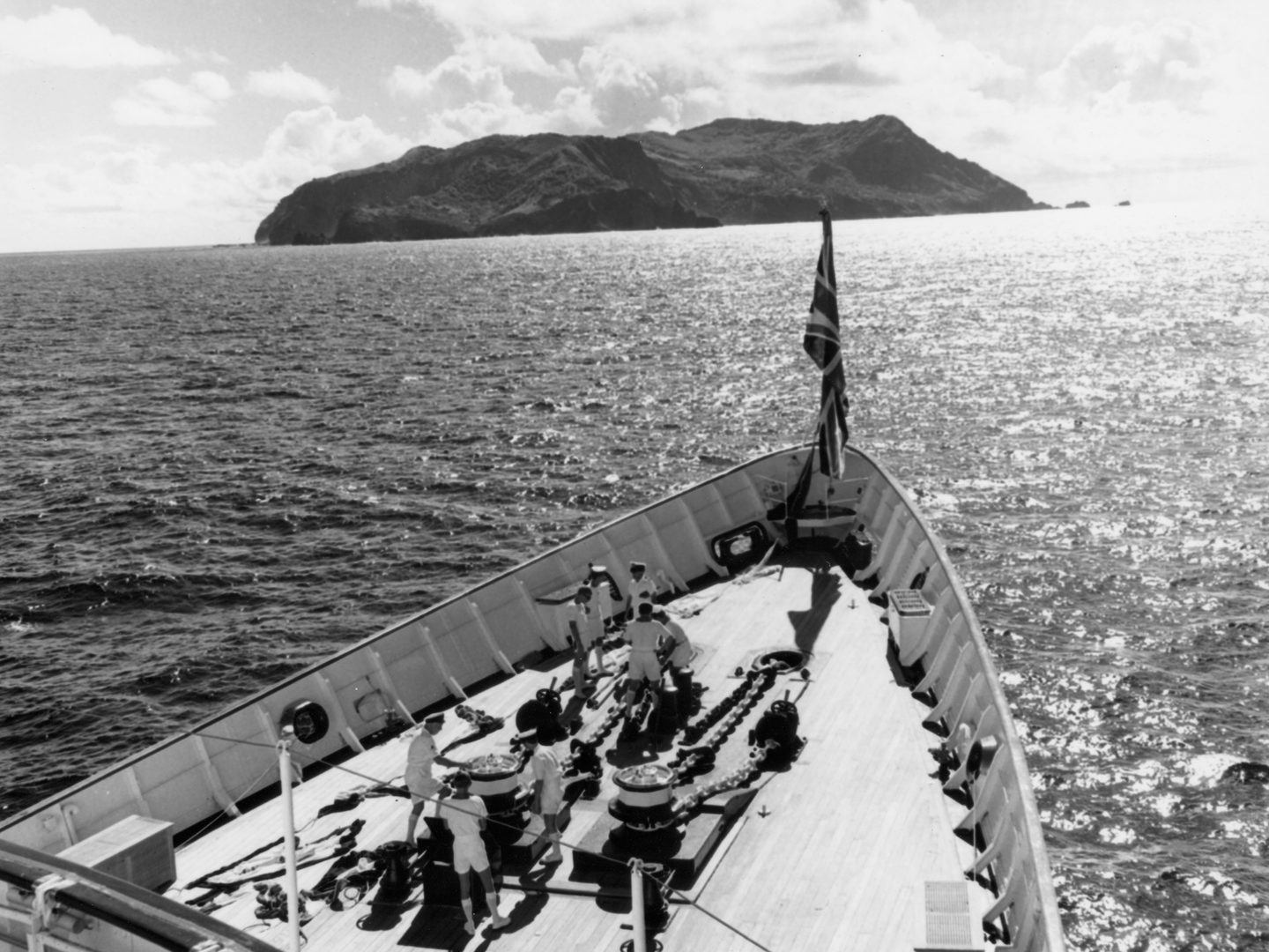 A historic black and white photo. In the foreground, 8 sailors in white uniforms are at work in the bow of the Royal Yacht Britannia. At the very front of the vessel is a hanging Union Flag. The island of Pitcairn is visible on the horizon.