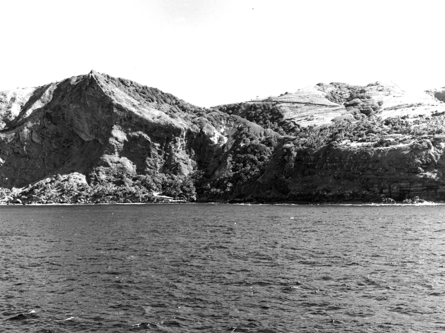 A historic black and white photo of Pitcairn taken from sea level. The sea is in the foreground, with the steep wooded slopes of Pitcairn rising out of the sea. The beginning of a road curving into the hills is just visible.