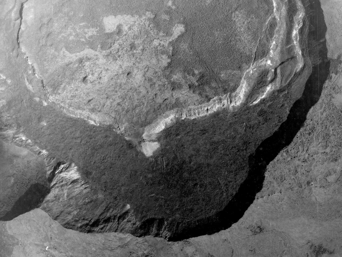 A black and white aerial photo of a forested landscape. A line of steep cliffs cast a thick band of shadow over a large crater. Opposite them is a second line of cliffs which show up white in the sun.