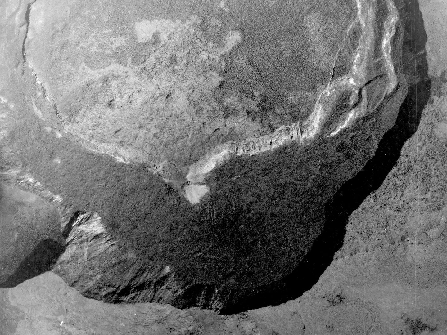 A black and white aerial photo of a forested landscape. A line of steep cliffs cast a thick band of shadow over a large crater. Opposite them is a second line of cliffs which show up white in the sun.