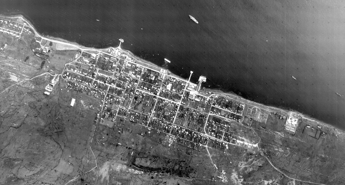 A historic black and white vertical aerial photo of a town on a coast. A couple of vessels are visible on the dark sea at the top of the image. The town is laid out along the coast in a grid, and has piers jutting out into the water.