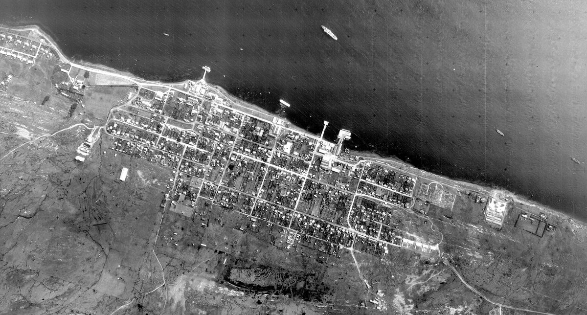 A historic black and white vertical aerial photo of a town on a coast. A couple of vessels are visible on the dark sea at the top of the image. The town is laid out along the coast in a grid, and has piers jutting out into the water.