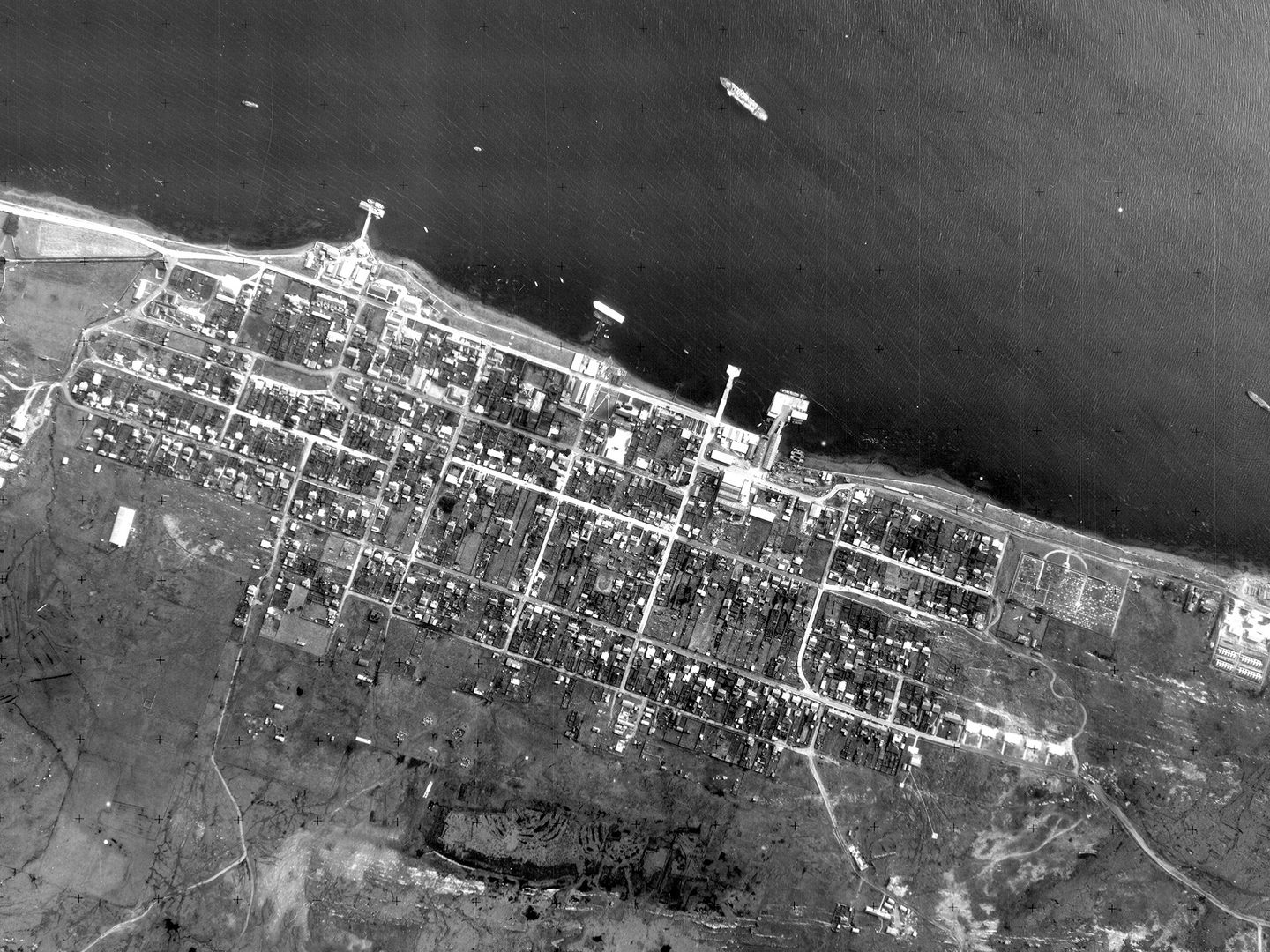 A historic black and white vertical aerial photo of a town on a coast. A couple of vessels are visible on the dark sea at the top of the image. The town is laid out along the coast in a grid, and has piers jutting out into the water.