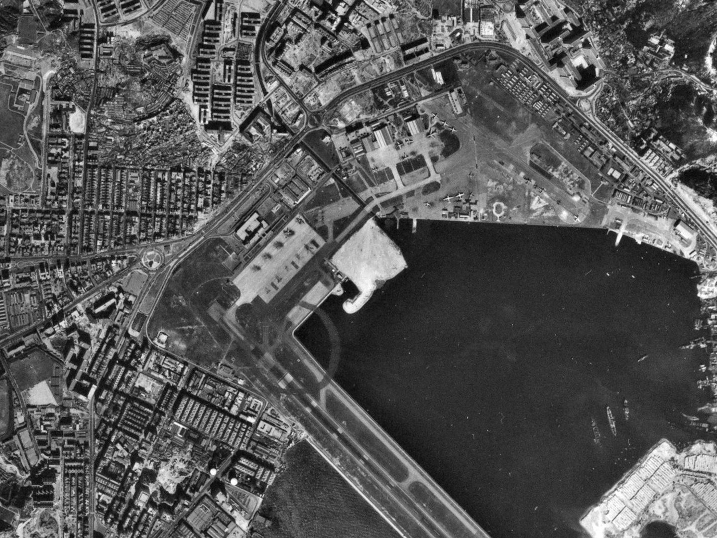 A black and white aerial photo of a built-up area of land, with the sea in the bottom right. Part of a runway projects over the sea. In the top left corner is an especially densely built-up area (Kowloon Walled City), surrounded by apartment blocks.