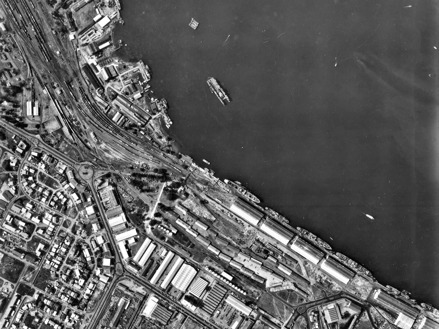 A black and white vertical aerial photo of a busy port area. The land is crowded with buildings railways and roads. Vessels of various descriptions are on the water and tied up at the quaysides.