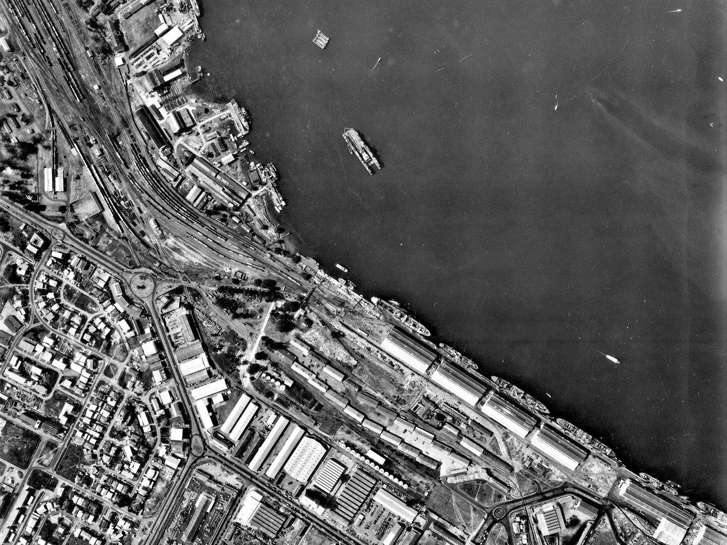A black and white vertical aerial photo of a busy port area. The land is crowded with buildings railways and roads. Vessels of various descriptions are on the water and tied up at the quaysides.