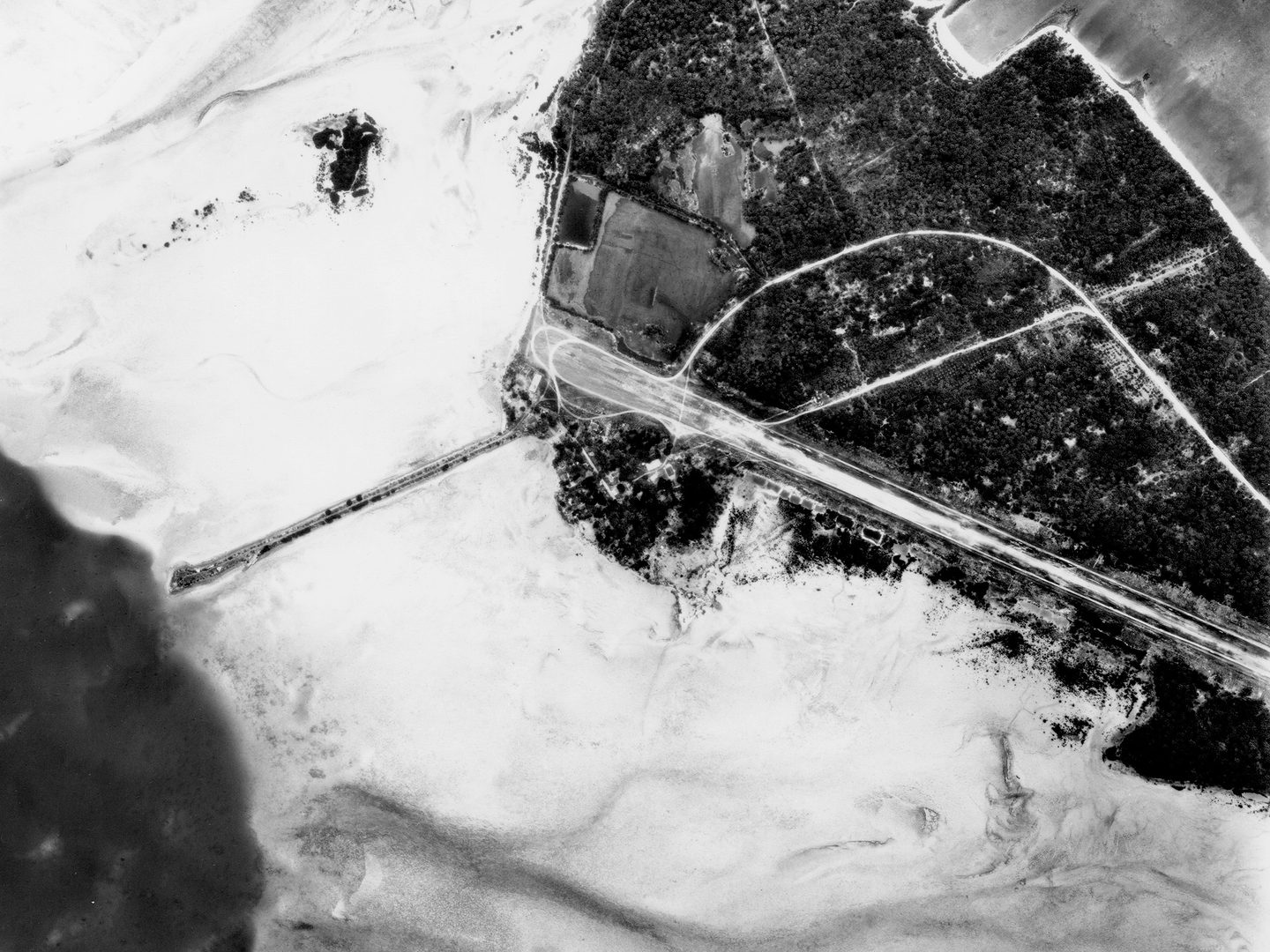 A historic black and white vertical aerial photo of an airport runway on an island. The island is rimmed with a sandy shoreline and surrounded by the sea.