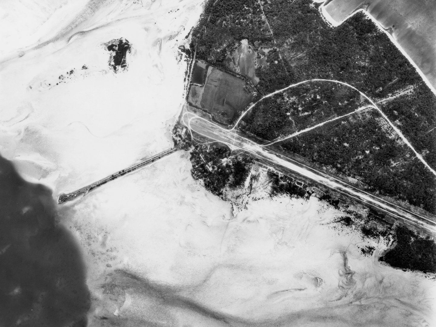 A historic black and white vertical aerial photo of an airport runway on an island. The island is rimmed with a sandy shoreline and surrounded by the sea.