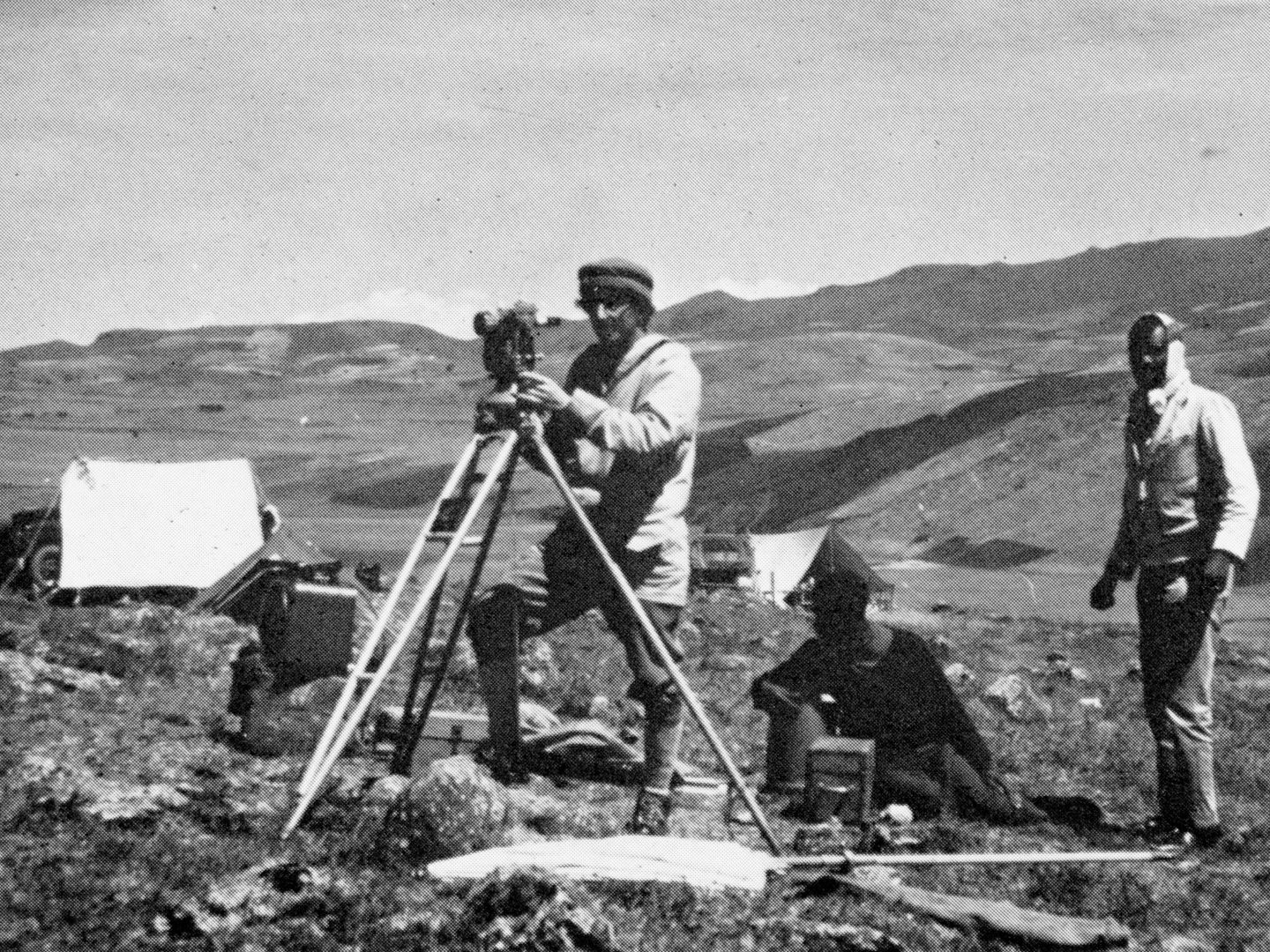 A black and white historic photo of 3 men surveying a hilly landscape in Ethiopia. A white man operates a piece of surveying equipment. Behind him are 2 black men, one sitting and one standing. White tents and hills are visible in the background.