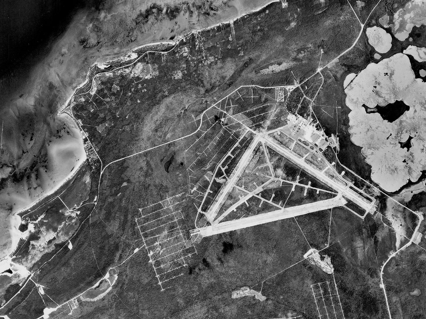 A historic black and white vertical aerial photo of an airfield on an island. A shoreline and the sea are visible in the top left corner.