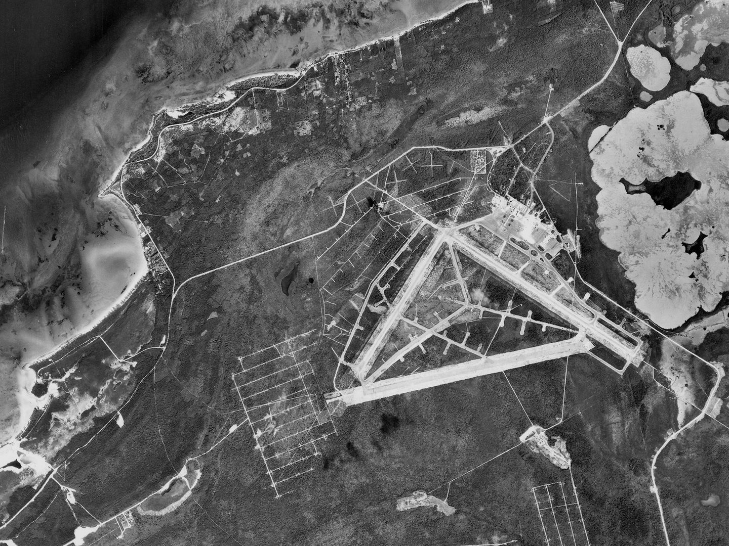 A historic black and white vertical aerial photo of an airfield on an island. A shoreline and the sea are visible in the top left corner.