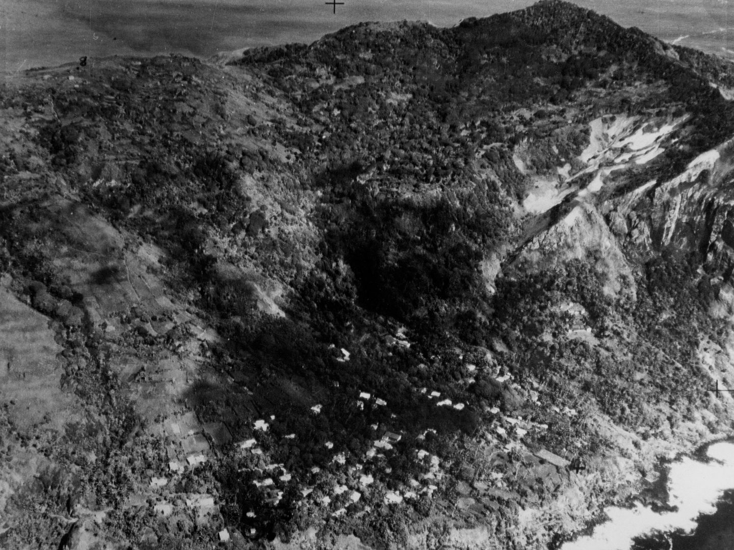 A historic black and white oblique aerial photo of the settlement of Adamstown on Pitcairn, with numerous white houses dotted throughout a wooded landscape. The settlement is just above the shore, with hills rising behind it.
