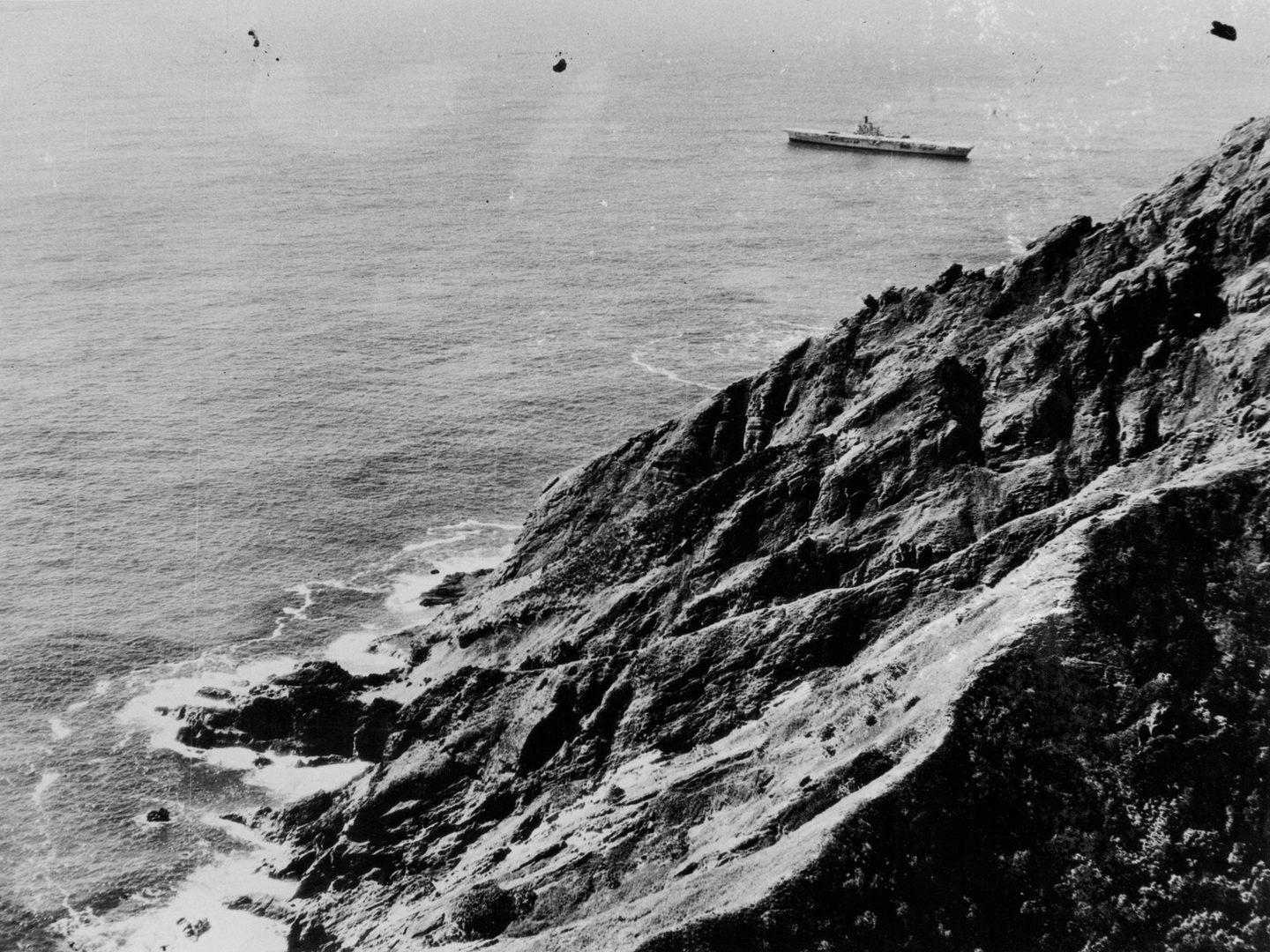 A historic black and white oblique aerial photo of the cliffs of Pitcairn dropping into the sea. In the distance is the aircraft carrier HMS Warrior.