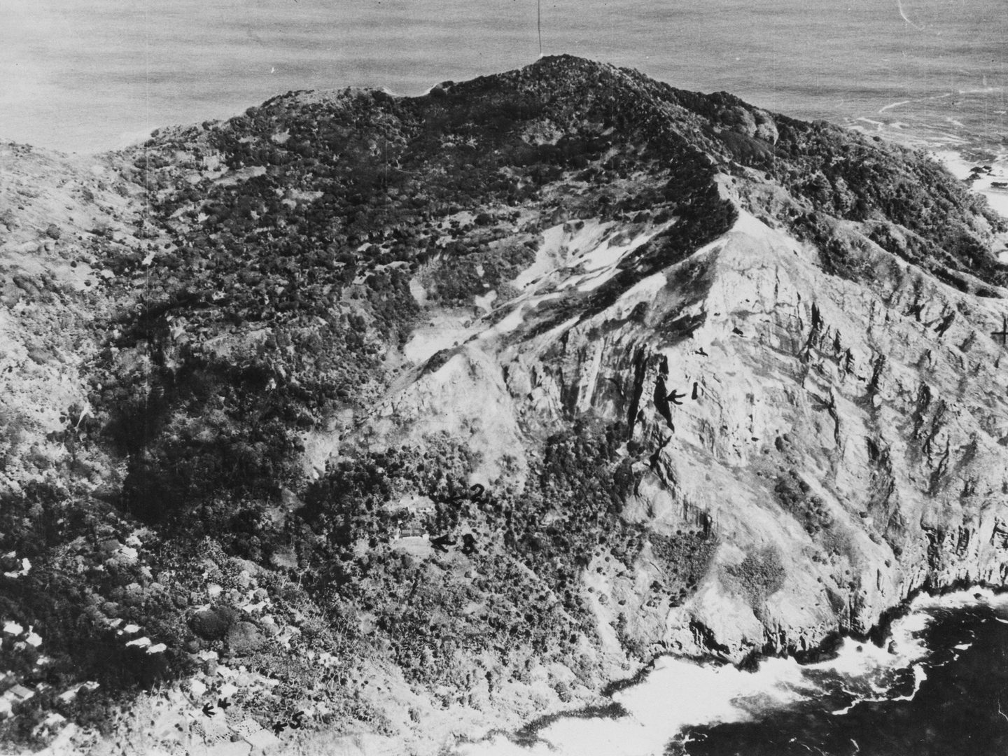 A historic black and white oblique aerial photo of Pitcairn, with cliffs and bare steep slopes rising to a mountainous peak. A small number of houses are visible among trees to the left.