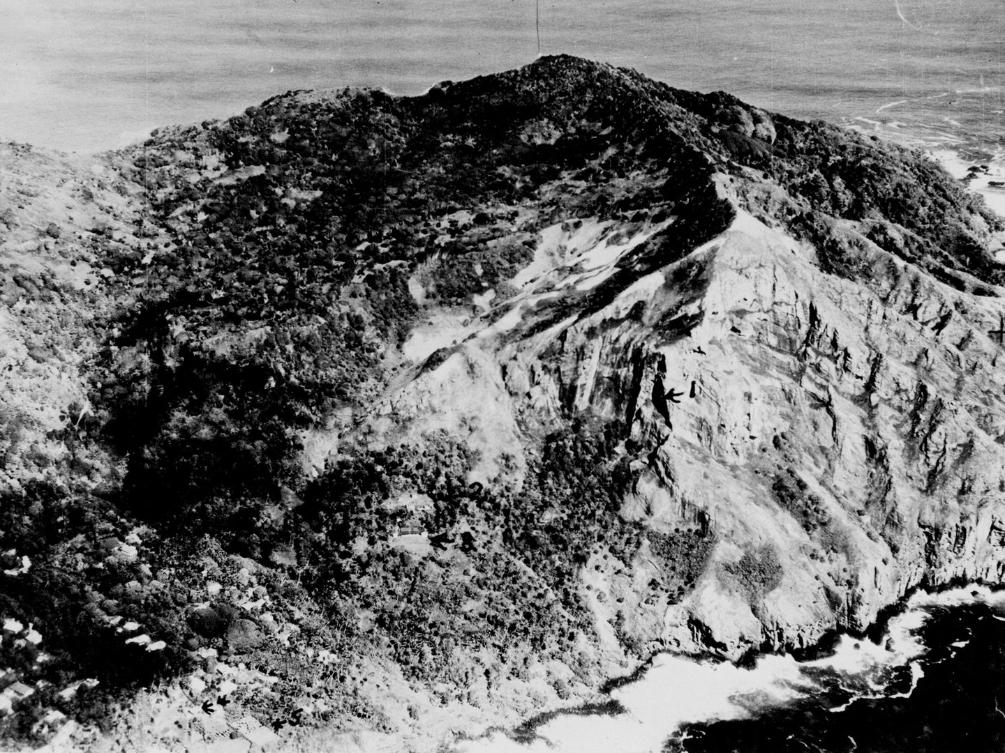 A historic black and white oblique aerial photo of Pitcairn, with cliffs and bare steep slopes rising to a mountainous peak. A small number of houses are visible among trees to the left.