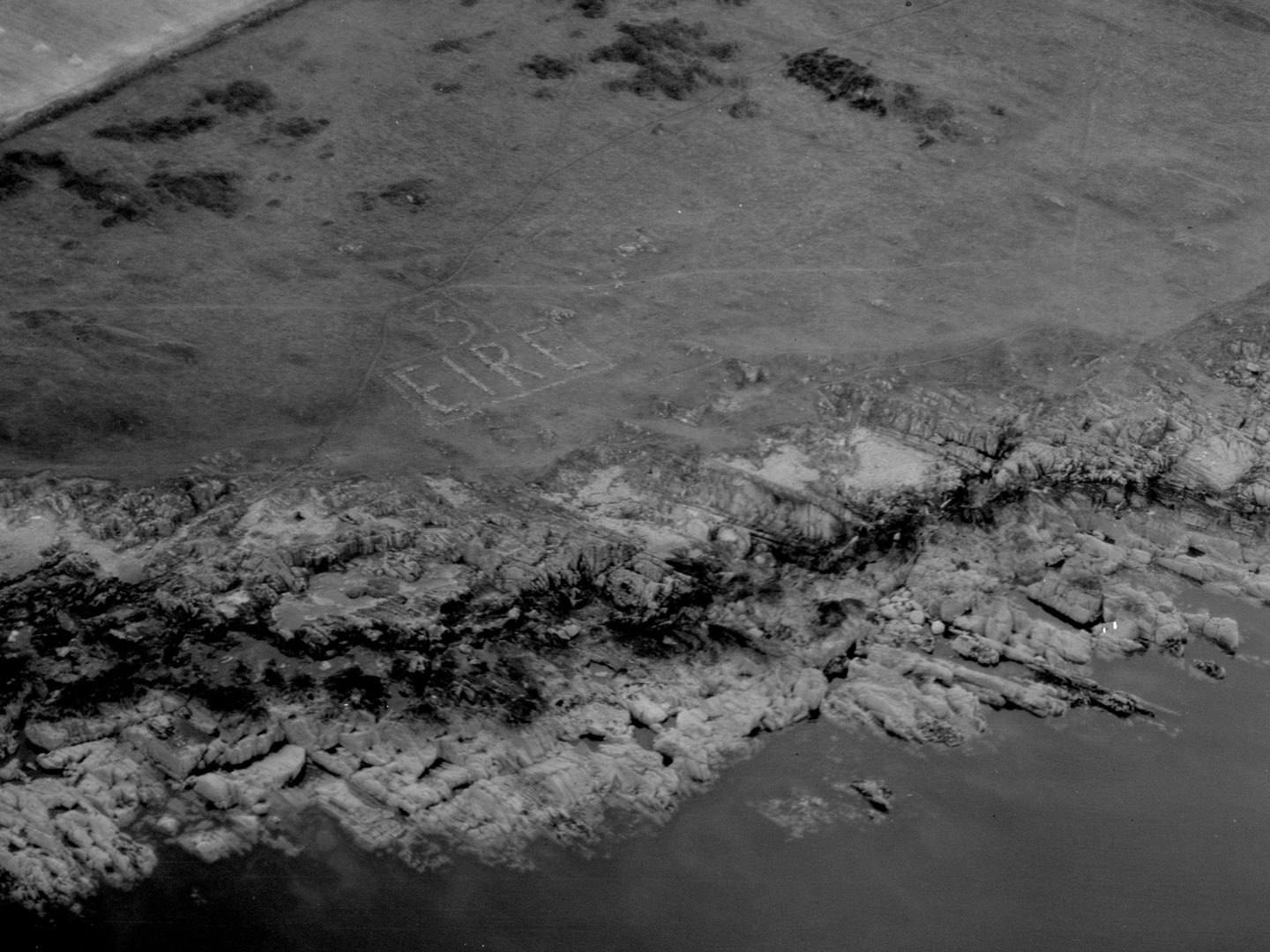 A historic black and white oblique aerial photo of a grassy field edged with a rocky shore that runs down to the sea. The letters '3 EIRE' are picked out in white on the grass.
